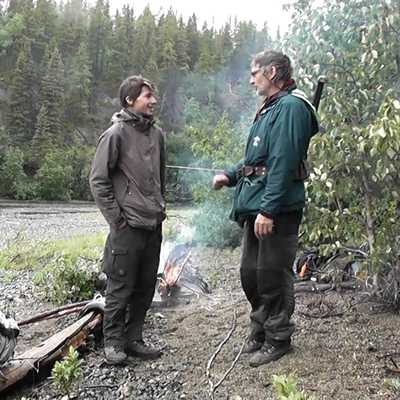 Norman Junker und sein Vater stehen im Gespräch an einem Lagerfeuer am Flussufer.