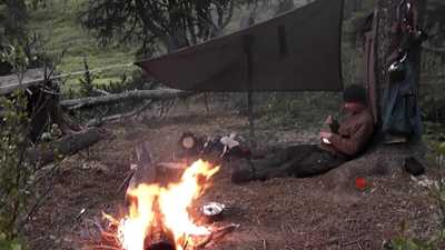 Lagerfeuer vor einem Tarp, dahinter ruht eine Person an einem Baum.