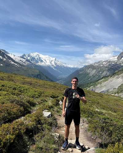 Norman Junker steht auf einem schmalen Bergpfad, dahinter Tal und schneebedeckte Gipfel.
